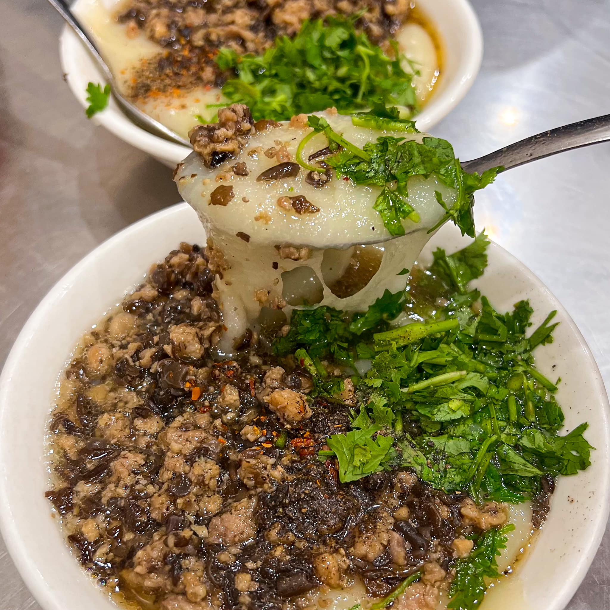 Close-up of a spoon lifting silky rice batter, minced pork, and crispy shallots from a bowl of bánh đúc nóng.