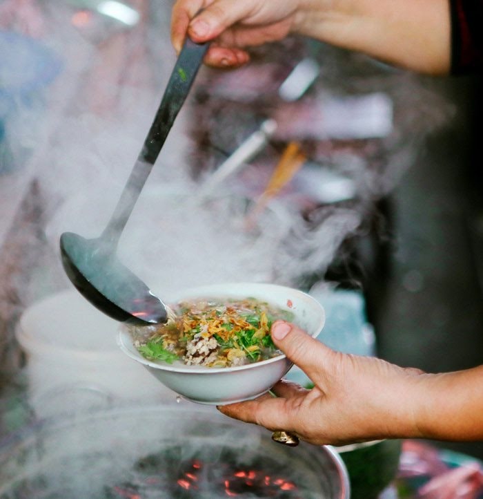 A person gently scooping a bowl of hot savory rice cake with fresh herbs and fish sauce broth in Hanoi.